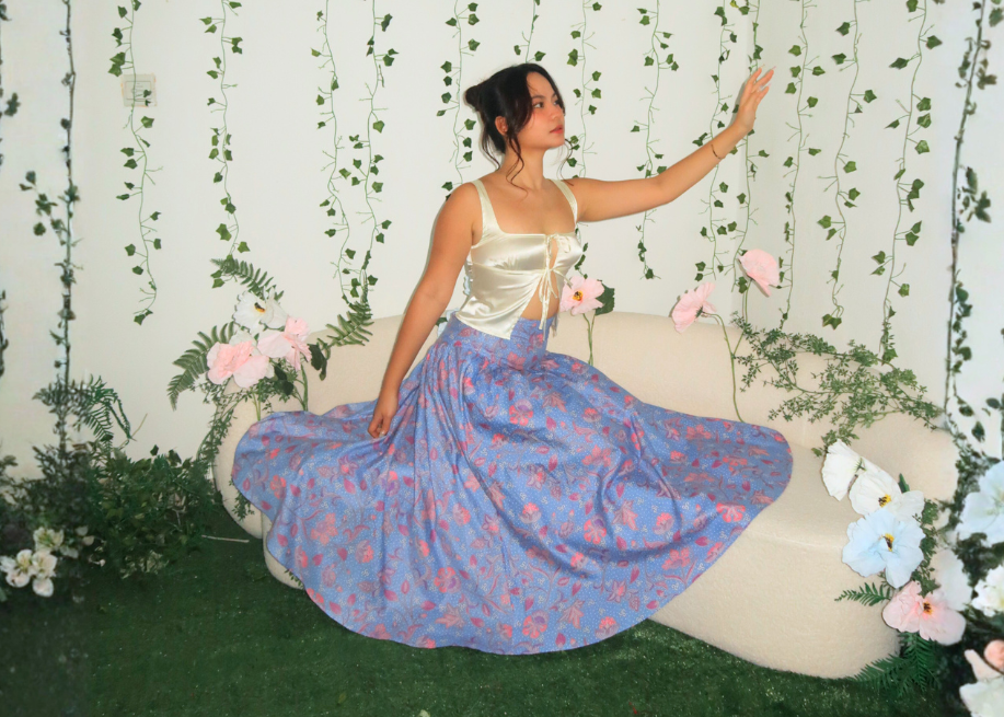 Woman in a blue skirt sitting on a white chair surrounded by greenery and flowers.